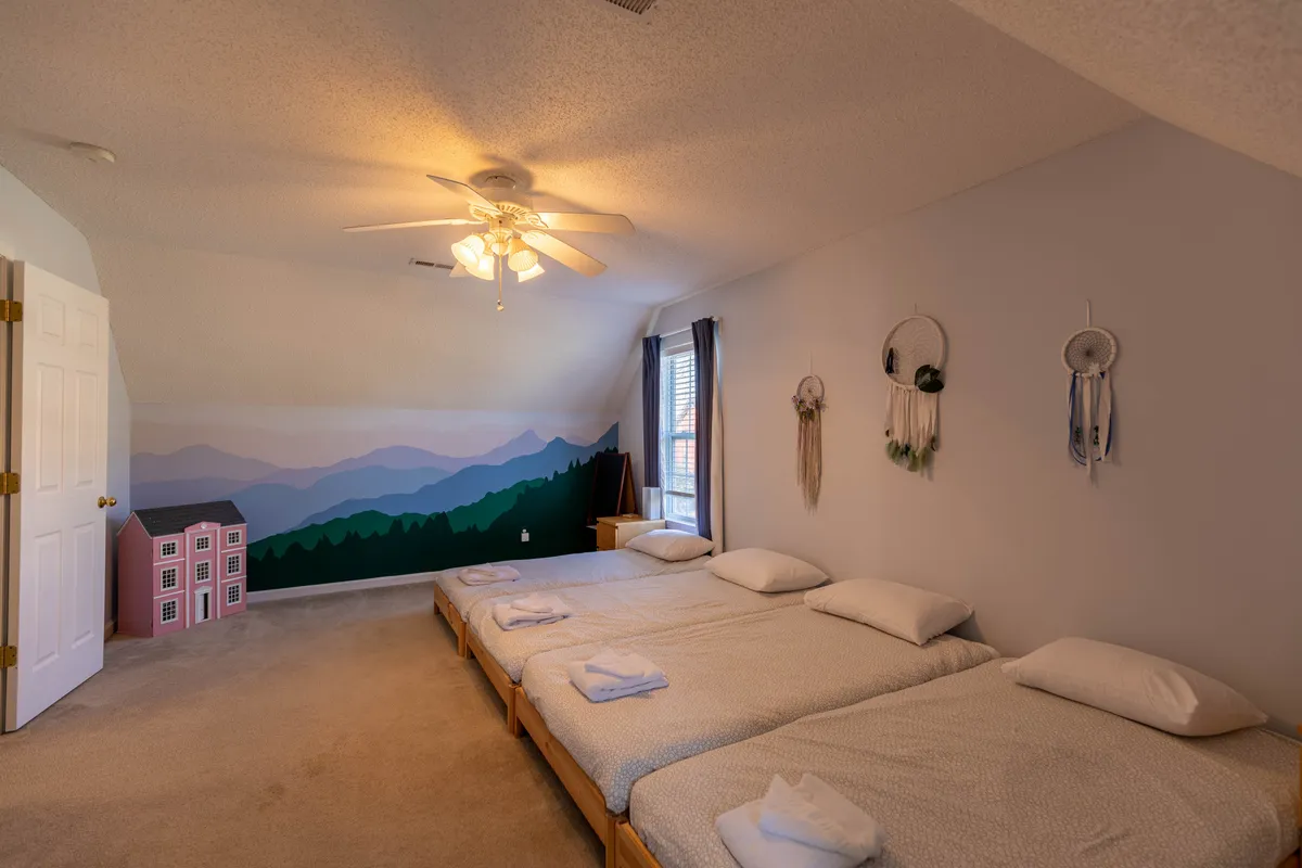 A cheerful bunk room with three single beds arranged in a row, a playful mountain mural on the wall, dreamcatchers by the window, and a pink dollhouse for added whimsy. The décor is clearly designed with children in mind, inviting shared moments and sleepovers.
