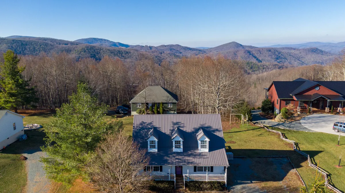 An exterior view showing the main house set against a backdrop of mountains, surrounded by trees and neighboring homes. The lawn offers space for gatherings, with scenic mountain vistas that invite exploration.