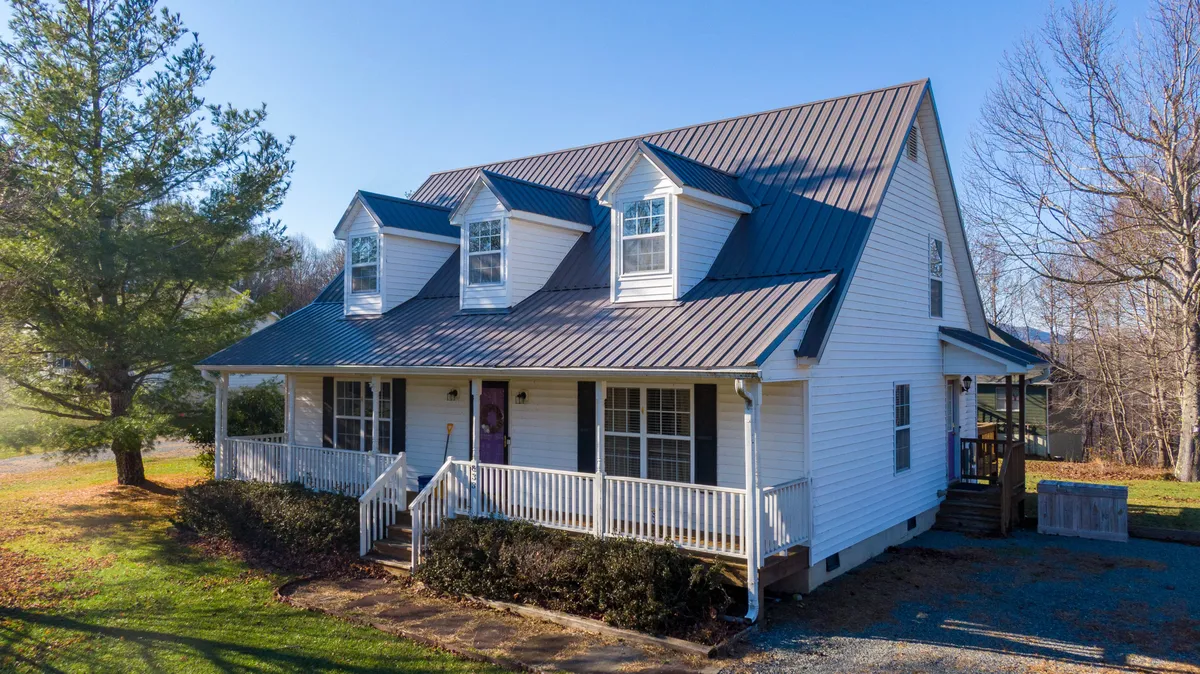 The hero image showcases the home's charming Cape Cod exterior, featuring a gable roof, expansive front porch with comfortable seating, and large windows glowing with warm interior light. The setting is inviting and framed by mountain scenery, promising sunset views and a welcoming space for morning coffee or festive gatherings.