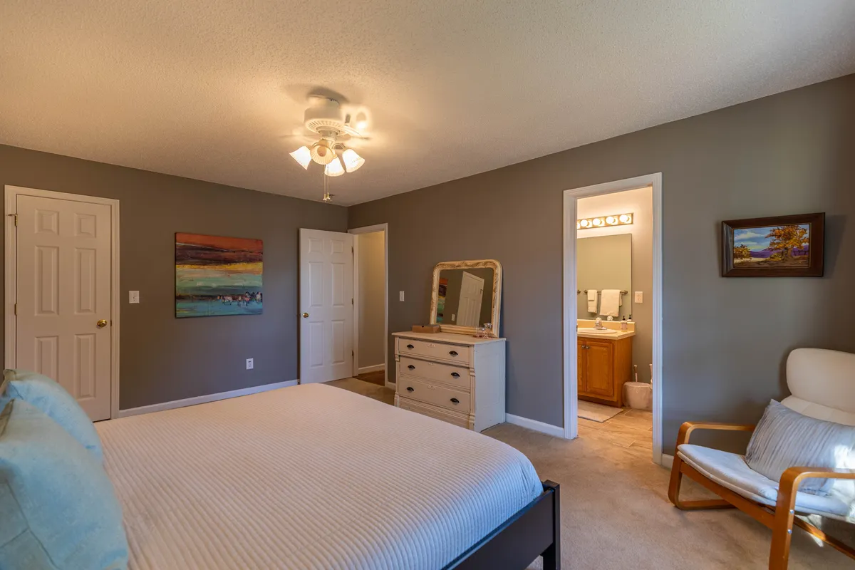 A calm, sunlit bedroom featuring a comfortable bed, a dresser, and a chair. Personal touches like artwork add warmth, and a door leads to an en suite bathroom, reinforcing the idea of privacy and convenience.