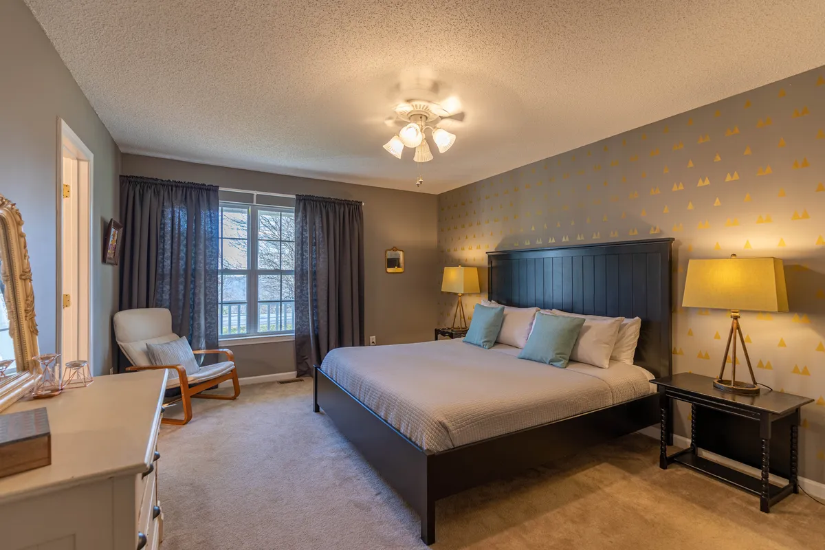 A serene king bedroom featuring a plush bed, soft linens, nightstands with decorative lamps, and an armchair by the window. Natural light fills the room, creating a peaceful and inviting atmosphere ideal for restful mornings.